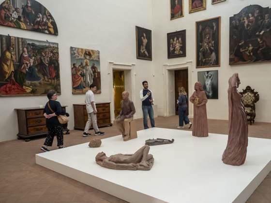 Cinque visitatori attraversano la sala mentre le sculture in terracotta restano ferme sulla pedana bianca: una figura distesa, una seduta, due in piedi. Alle pareti le pale antiche, luce naturale dall'alto.