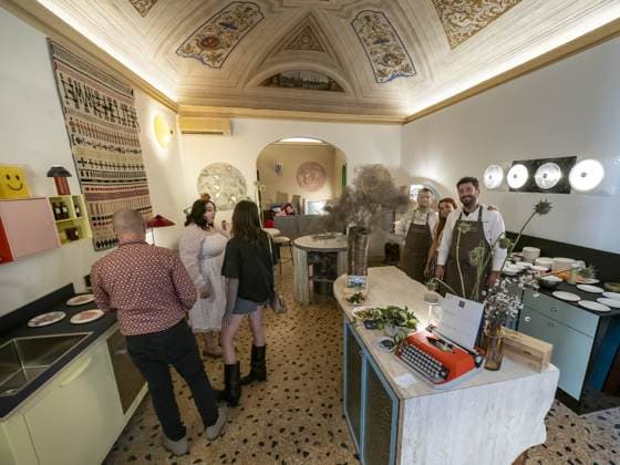 Quattro persone in grembiule posano dietro il bancone di marmo, sotto il soffitto affrescato; a sinistra tre visitatori si fermano a guardare i piatti in fila. La sala è ancora tranquilla, il servizio deve cominciare.