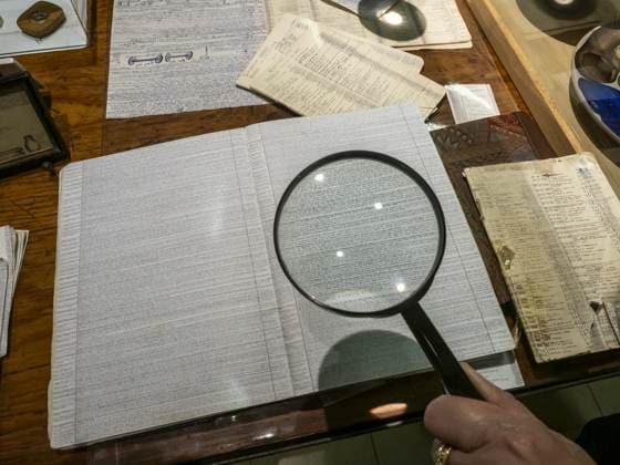 Una mano tiene la lente d'ingrandimento sopra la pagina fitta di un registro; intorno, sul piano di legno, altri quaderni rilegati e un disegno tecnico. Si legge una riga alla volta.