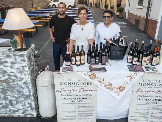 Tre persone dietro il banco della cantina, le bottiglie schierate su più file e il secchiello del ghiaccio al centro. Alle loro spalle i tavoli in strada sono ancora vuoti.