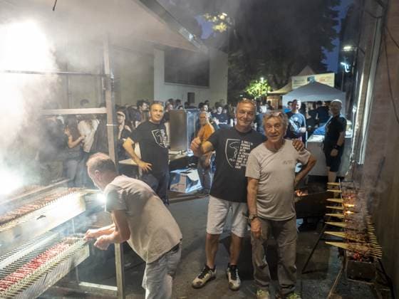 Uno si china a girare gli spiedini sulla griglia lunga, gli altri tre si voltano verso l'obiettivo; il fumo prende metà dell'inquadratura. Dietro, la fila di gente al gazebo e il cielo ancora blu.
