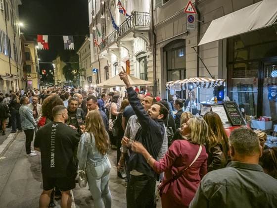 La via è piena da un lato all'altro: un uomo alza il bicchiere sopra la testa mentre il chiosco a destra continua a servire sotto la tenda a righe. È già notte, i drappi pendono dai palazzi.