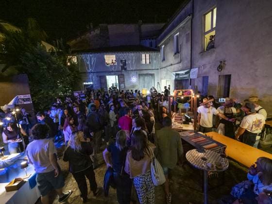 Dall'alto il cortile è pieno: un centinaio di persone in piedi fra i banchi e il tavolone di legno addossato al muro, sotto lo striscione «Cucina del Condominio». Notte piena, qualcuno segue la scena dalla finestra illuminata.